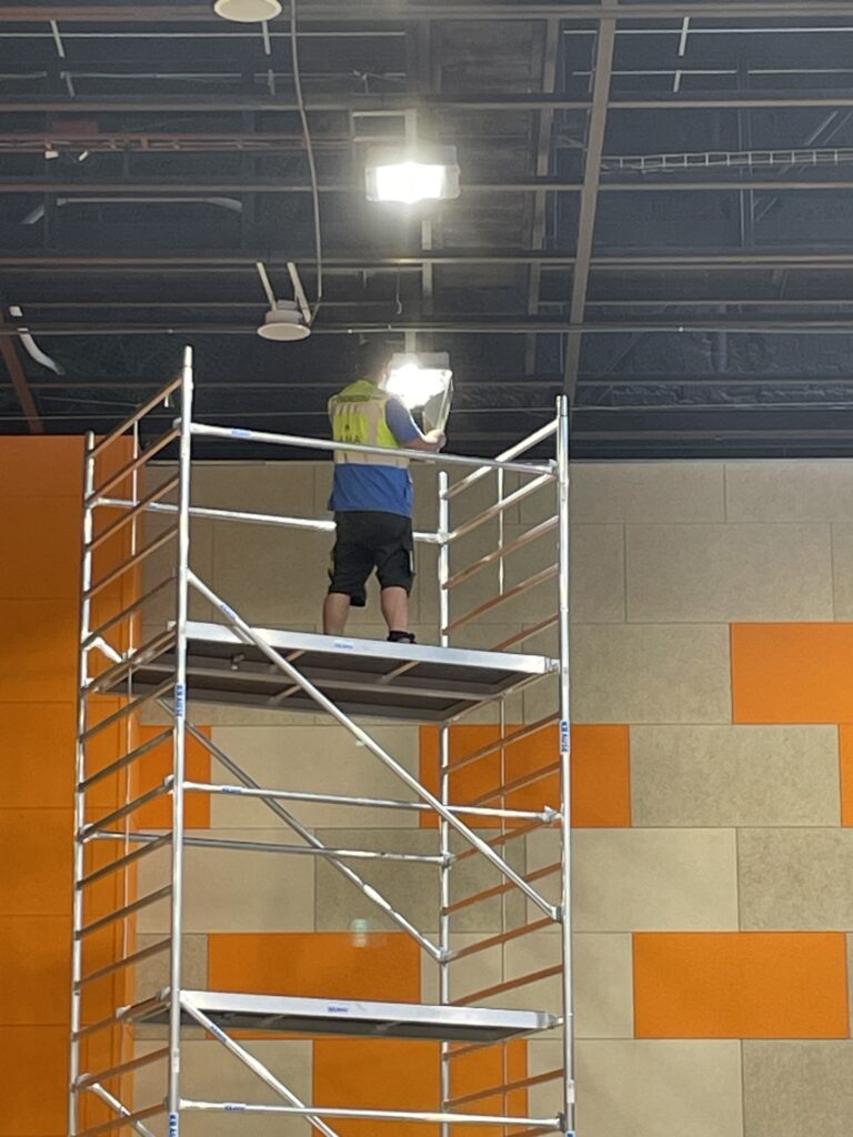 Pracovník ve sportovní hale při výměně osvětlení na lešení